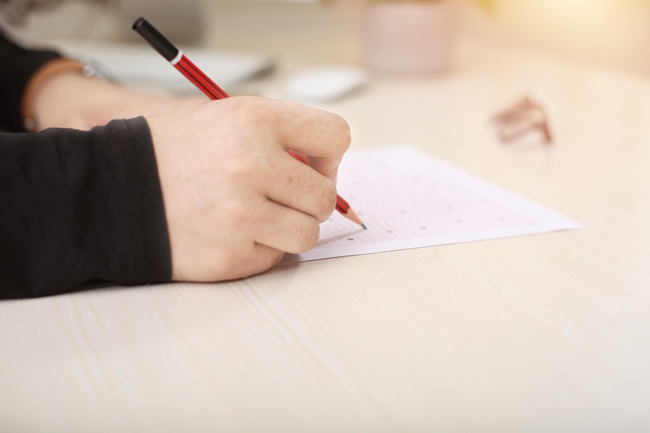 Osoba pisząca czerwonym długopisem na kartce papieru, siedząca przy jasnym biurku. W tle widoczne są rozmazane przedmioty, takie jak okulary i kubek, co sugeruje spokojne, skupione środowisko pracy lub nauki.